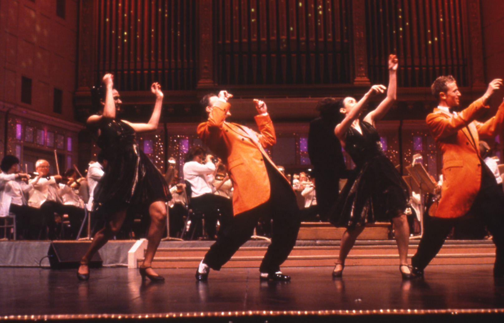 Two male dances (in orange 1950s style jackets) and two female dancers in dresses dance with arms raised in a line across Symphony Hall stage