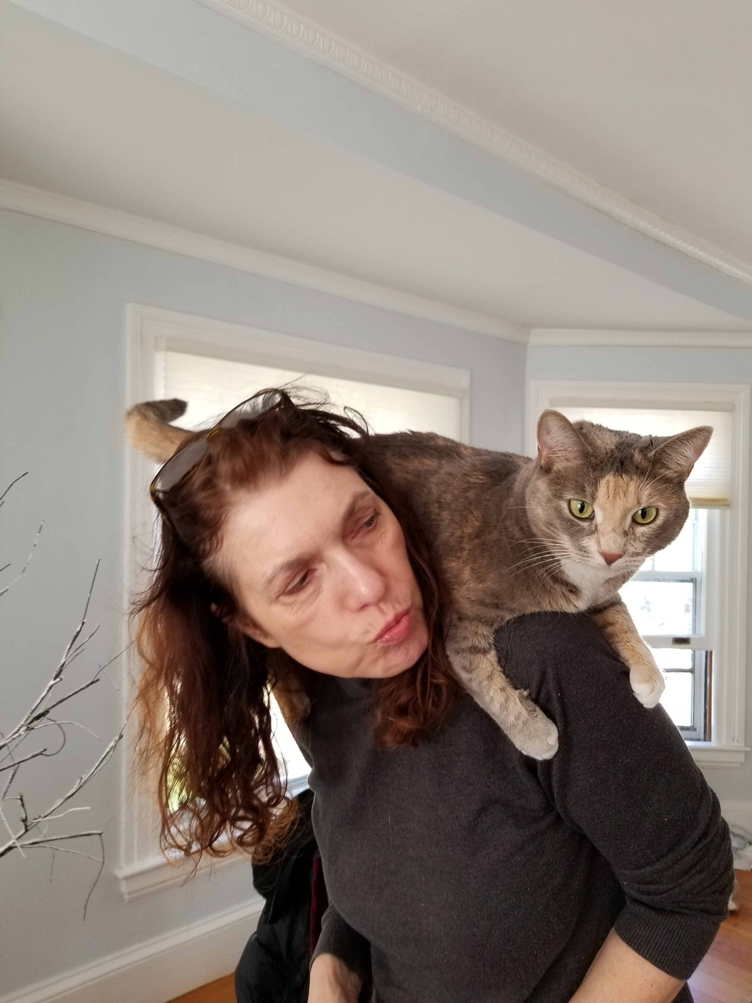 A woman wearing a black sweater stands with a brown cat sitting on her shoulders