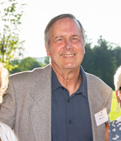 A man wearing a gray jacket smiles while standing outside in front of trees