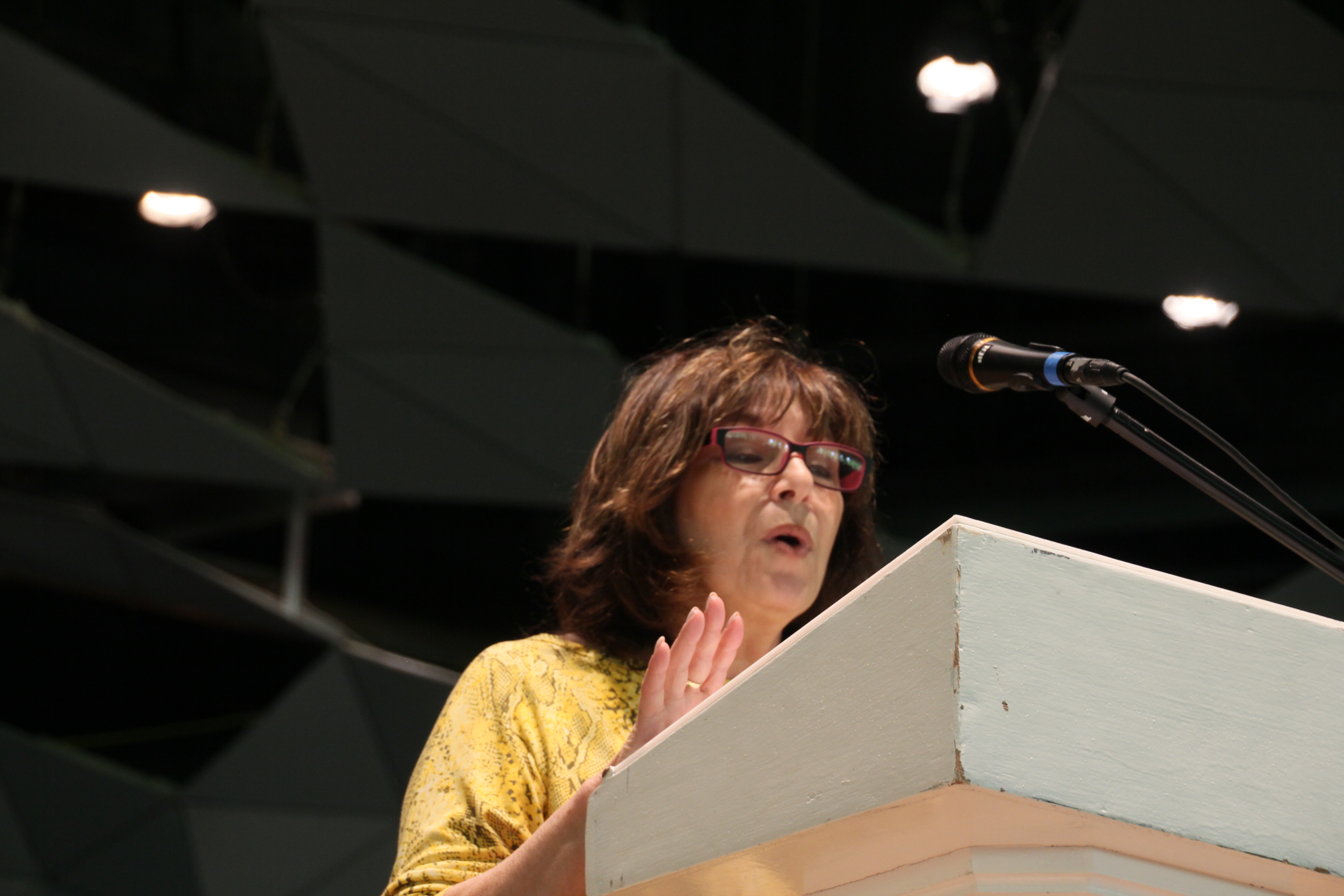 A woman wearing glasses and a yellow shirt stands at a podium with a microphone, in the middle of speaking, a dark background behind her