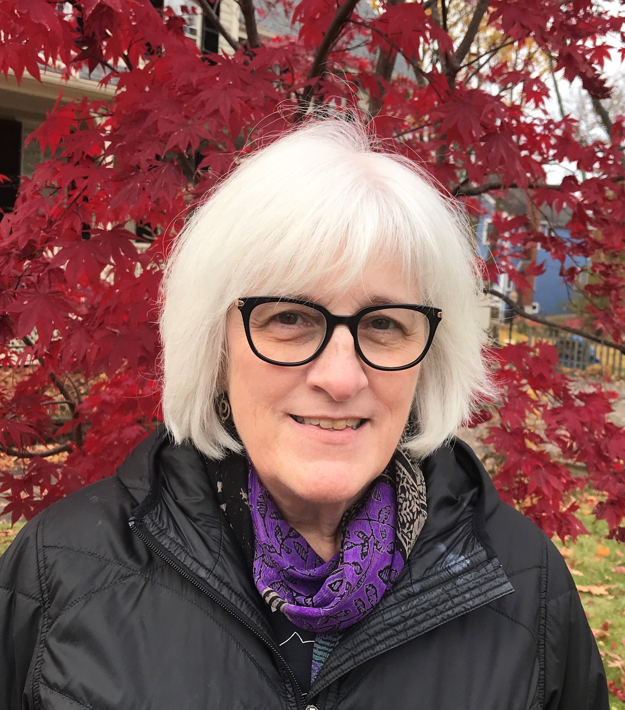 A woman wearing a black jacket and purple scarf stands in front of a tree with red leaves