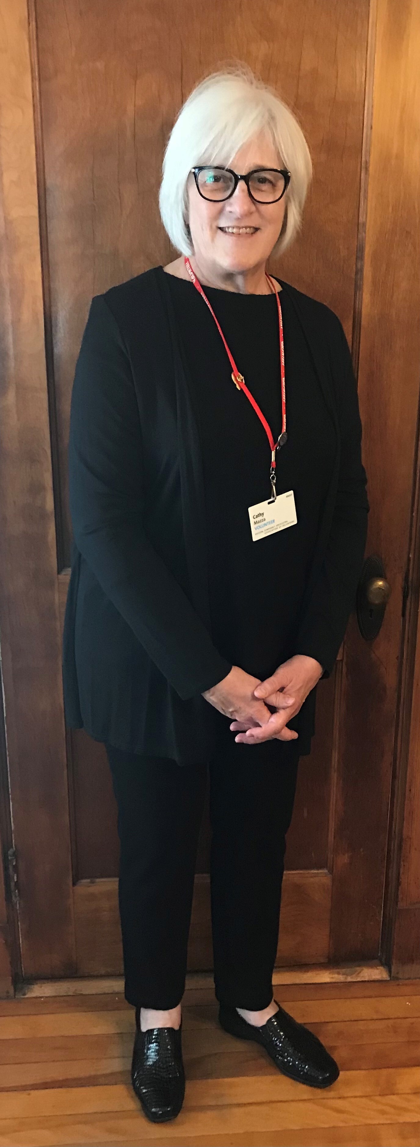 Woman wearing matching black long-sleeve shirt and black pants with a red lanyard standing in front of a  wood-paneled wall