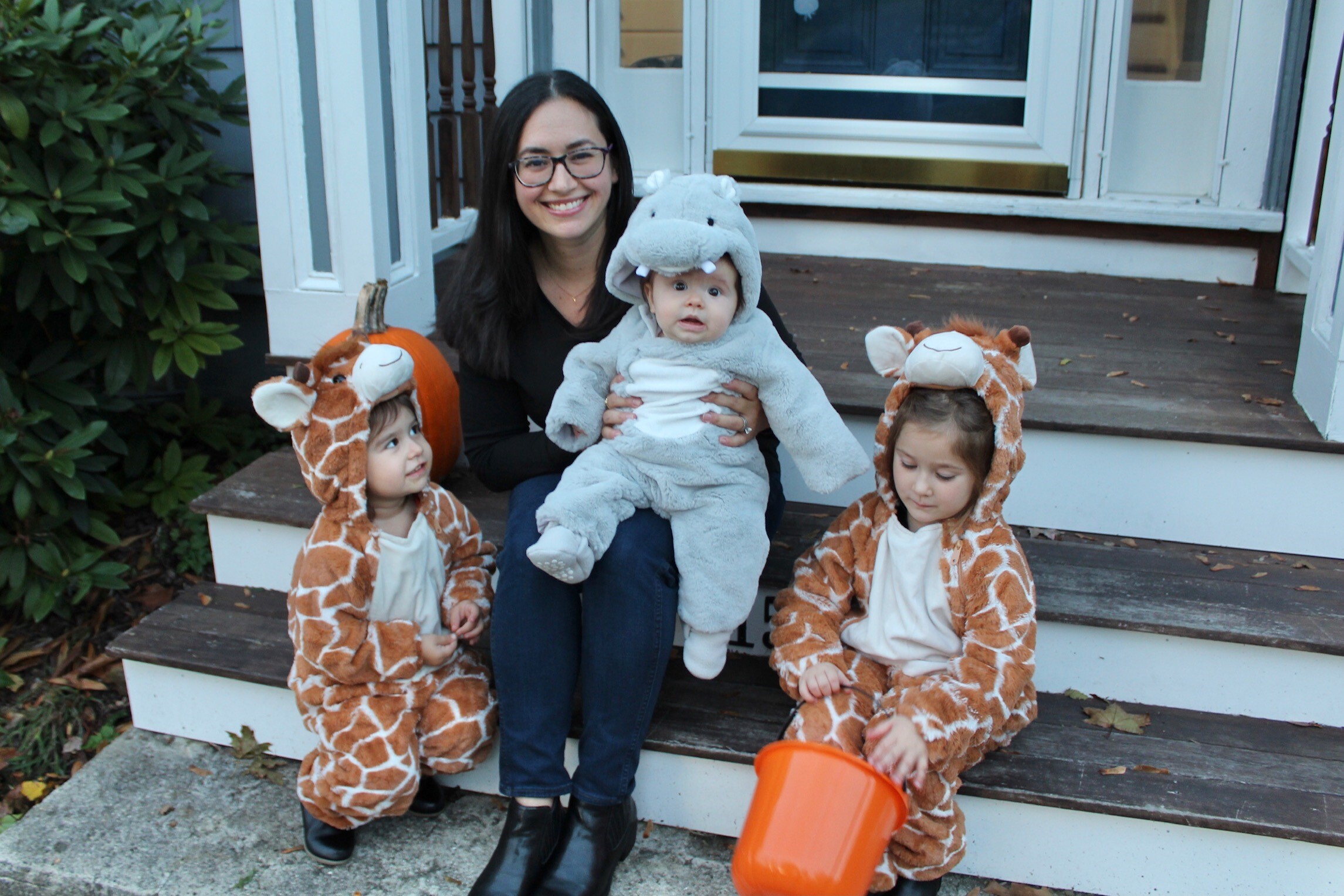 An adult woman sits on the front steps of a house with two children dressed as giraffes and a baby dressed as a hippopotamus