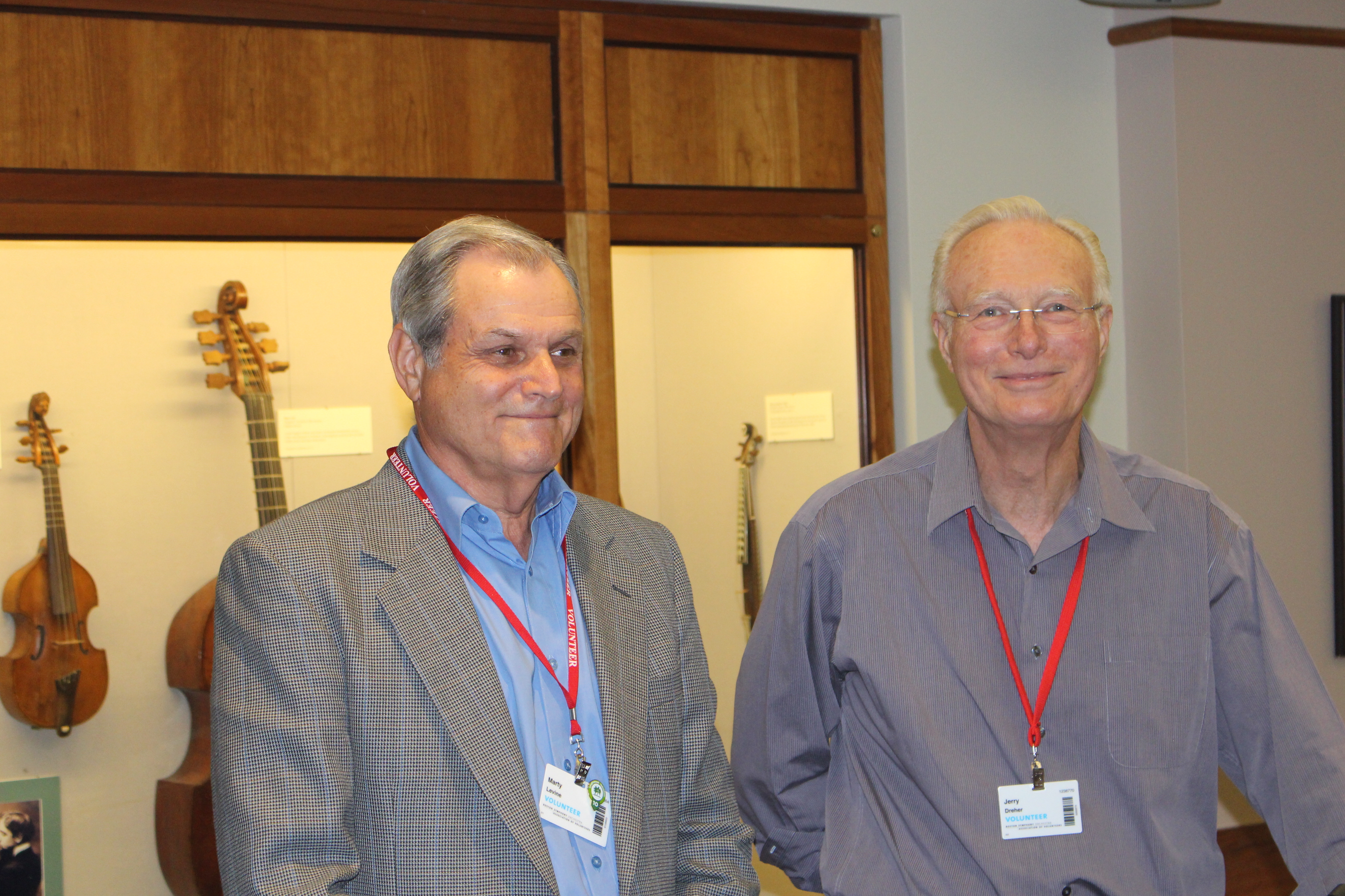 Two men, the left wearing a gray sportcoat and a blue shirt and the right wearing a gray button-down shirt, smiling in front of a display of  musical instruments