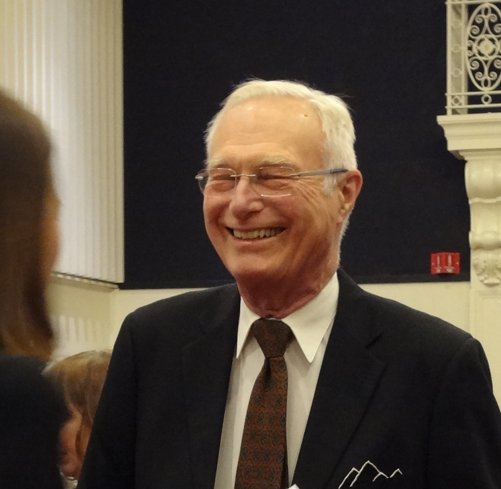 Smiling man with white hair and glasses wearing a suit with a brown tie