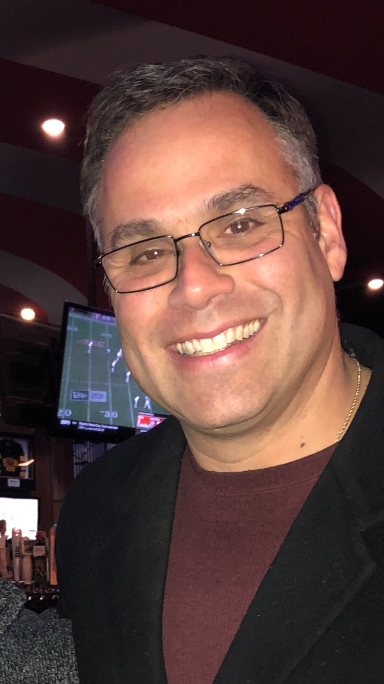 Man wearing glasses and a black coat over a maroon shirt smiling