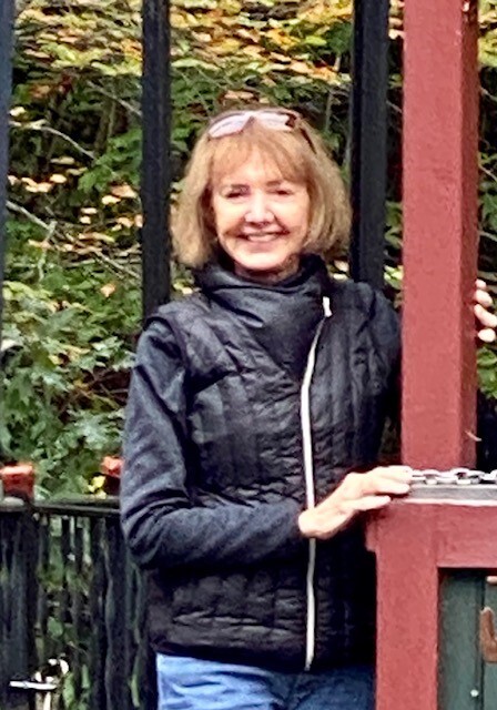 A woman wearing a black vest smiles as she stands in front of some trees