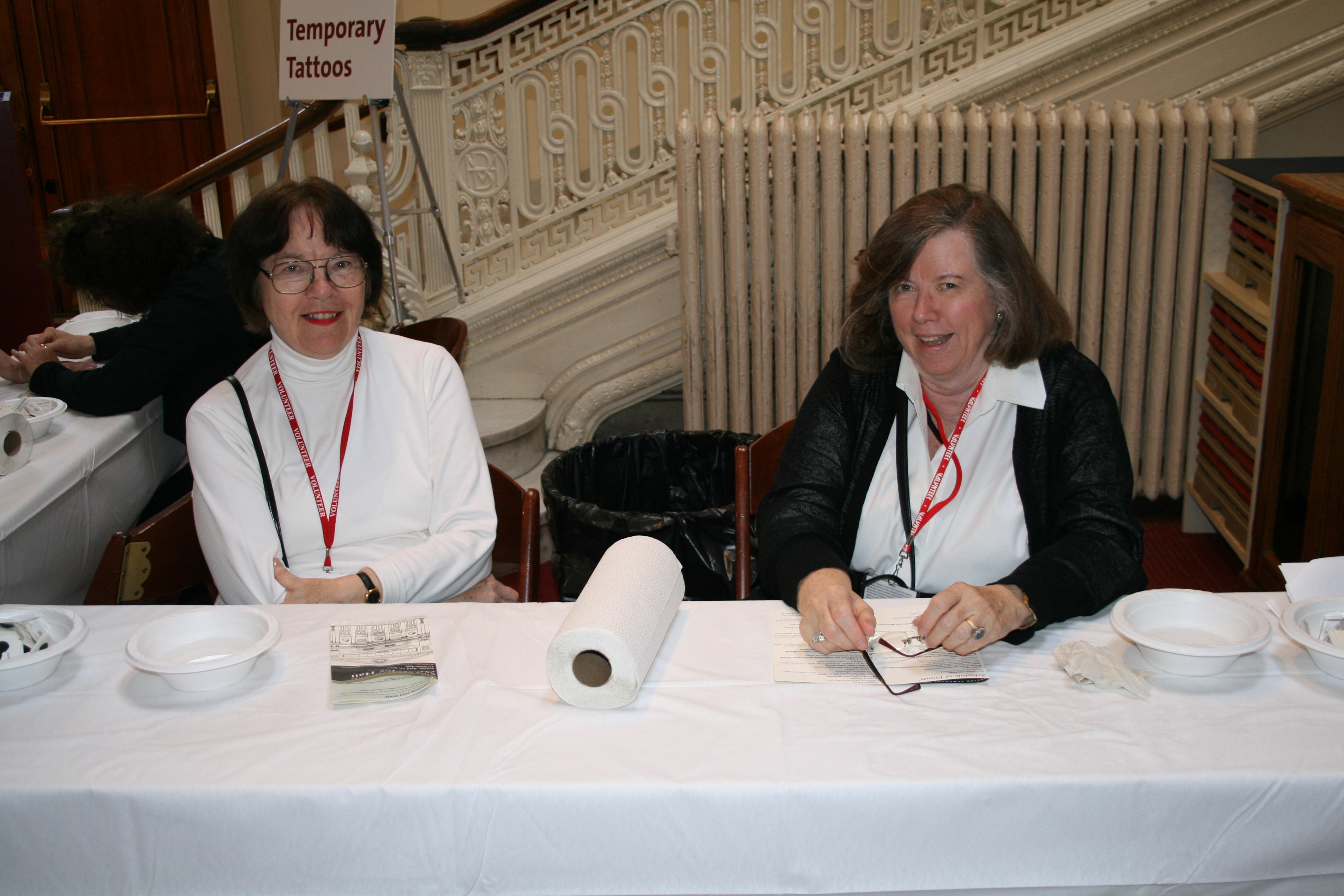 Two woman, one with black hair and glasses and wearing a white shirt, the other with brown hair and wearing a black jacket over a white shirt, smile while seated at a table with a white tablecloth