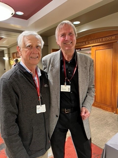 Two men, one wearing a gray suitcoat over a black shirt and pants, one wearing a gray pull-over sweater and glasses, stand together in front of a wood-paneled background inside a room.