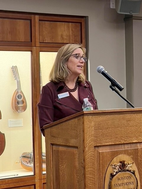 A woman with light hair and wearing glasses and a purple blouse stands at a podium, speaking into a microphone, in front of a lit cabinet displaying musical instruments