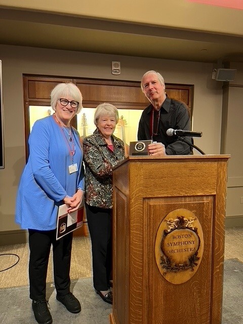 A shorter woman wearing a gray shirt and black pants stands behind a podium between a woman wearing a light blue blouse and black pants and a man wearing a black shirt, in front of a backlit cabinet