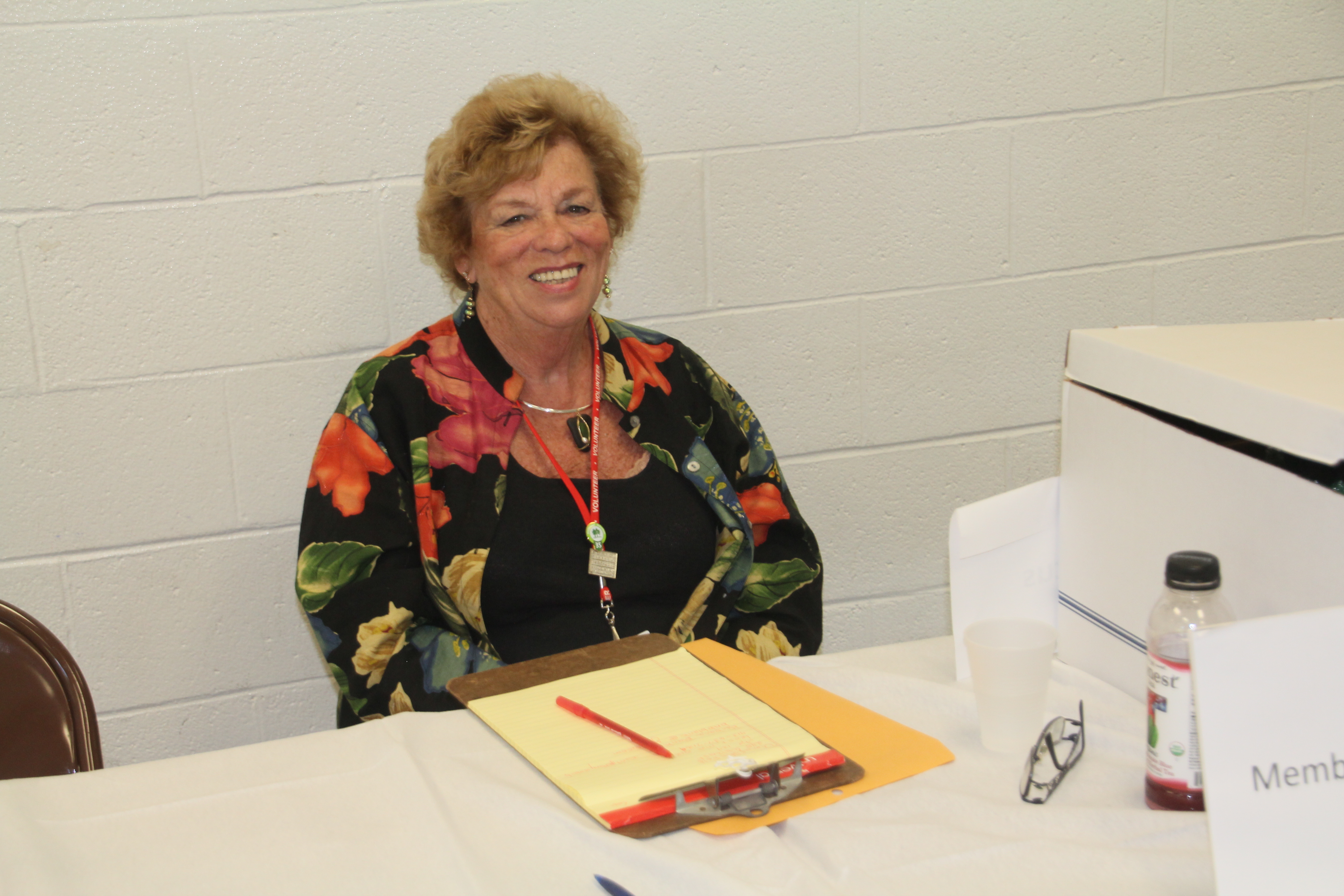 Woman with brown hair and wearing a black shirt with floral patterns sits at a table in front of a clip board in front of a white wall