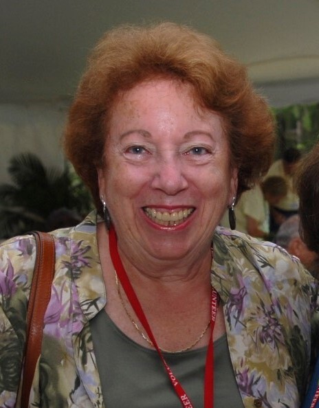 Woman with red hair and wearing a green floral print shirt and a red lanyard, smiling while standing inside a tent in front of others