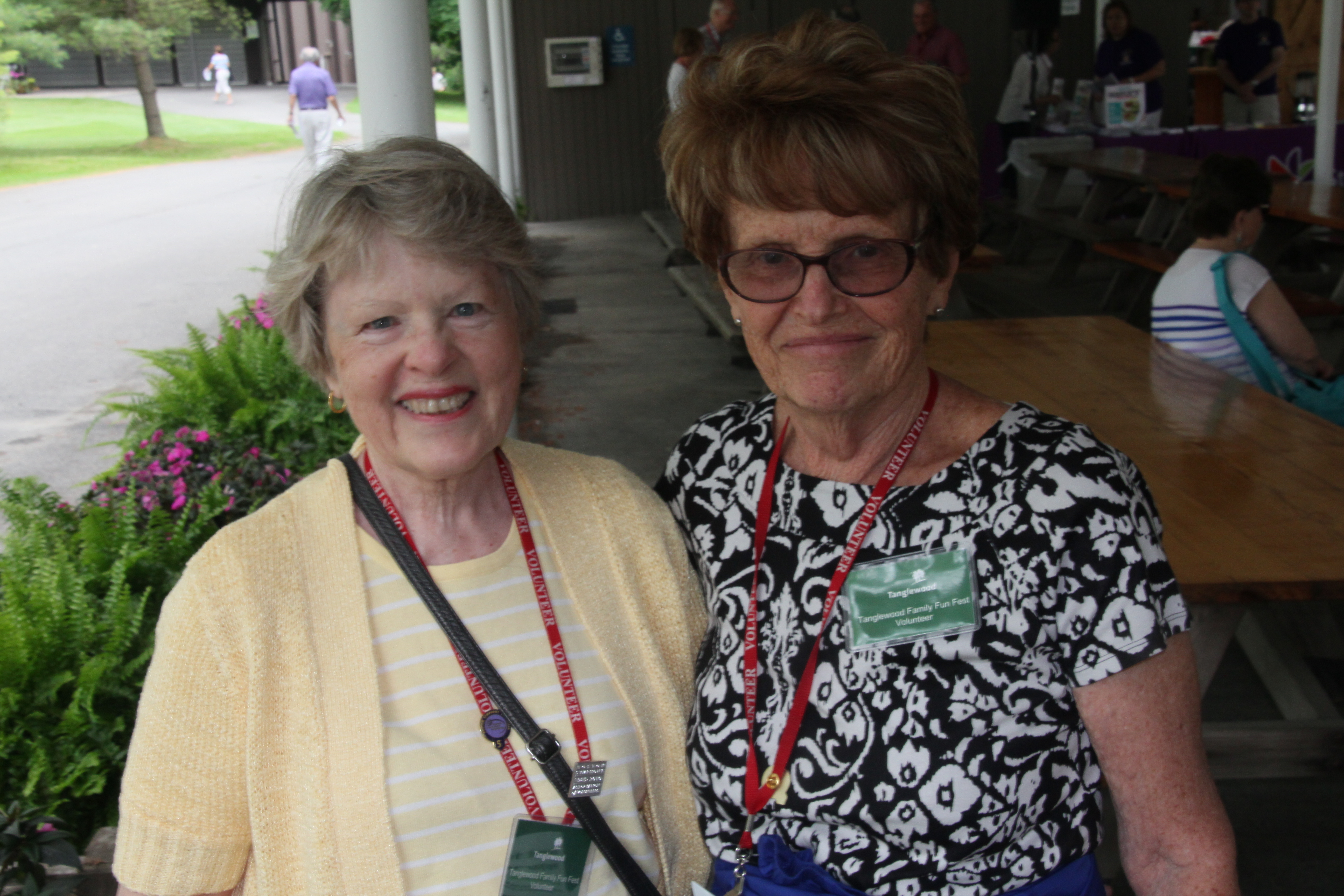 Two women, one wearing a yellow shit on the left and one wearing glasses and a black and white print shirt, smiling together outdoors