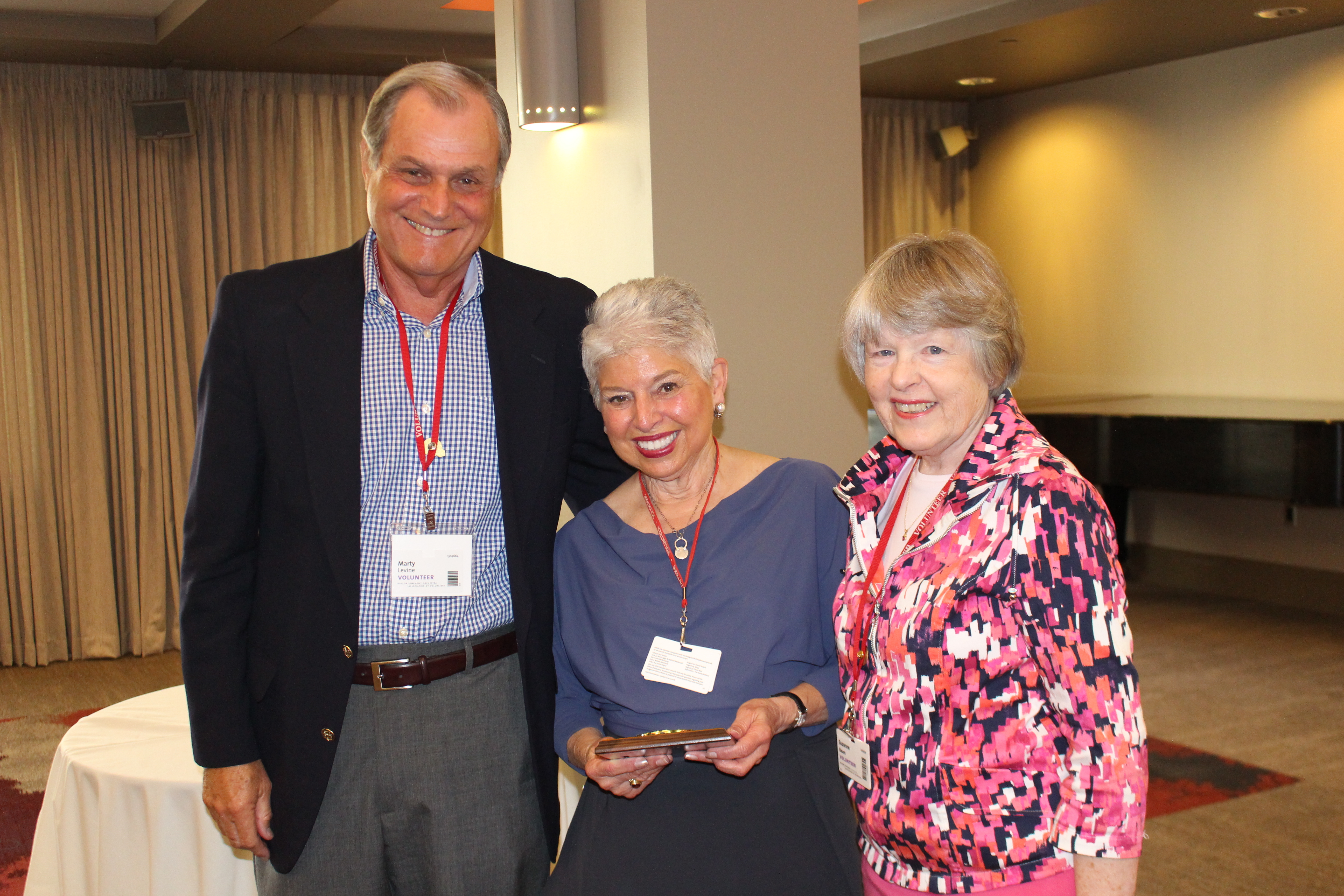 Tall man wearing sportcoat and lanyard stands with woman wearing blue shirt and holding an award and another woman wearing a pink floral shirt, smiling together