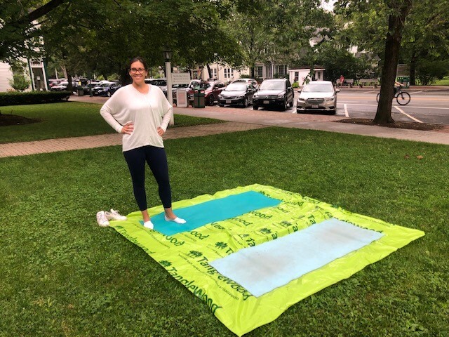 A woman stands on a green tarp on some grass with two yoga mats on the tarp