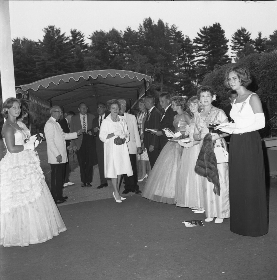 A black-and-white photo of women wearing gowns and men wearing suits standing on either side of the opening to a canopy covering a walkway, the tops of trees seen behind them