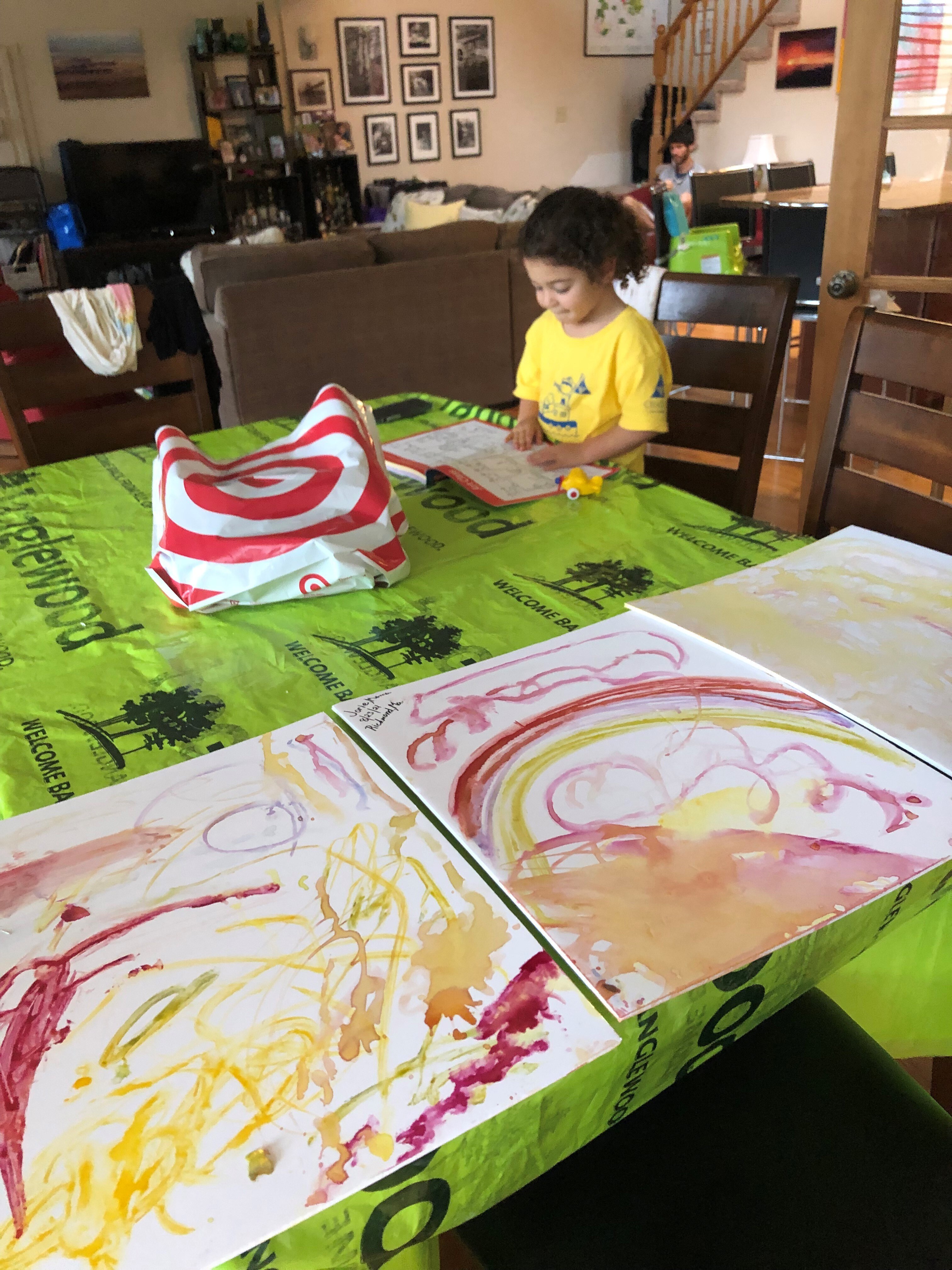A little girl reads a book by three finger paintings sitting on a green tarp