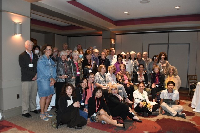 A group of people, many middle-aged or older, stand and sit in a group together to pose for the camera, inside a room with white walls and ceilings with wooden accents and trim