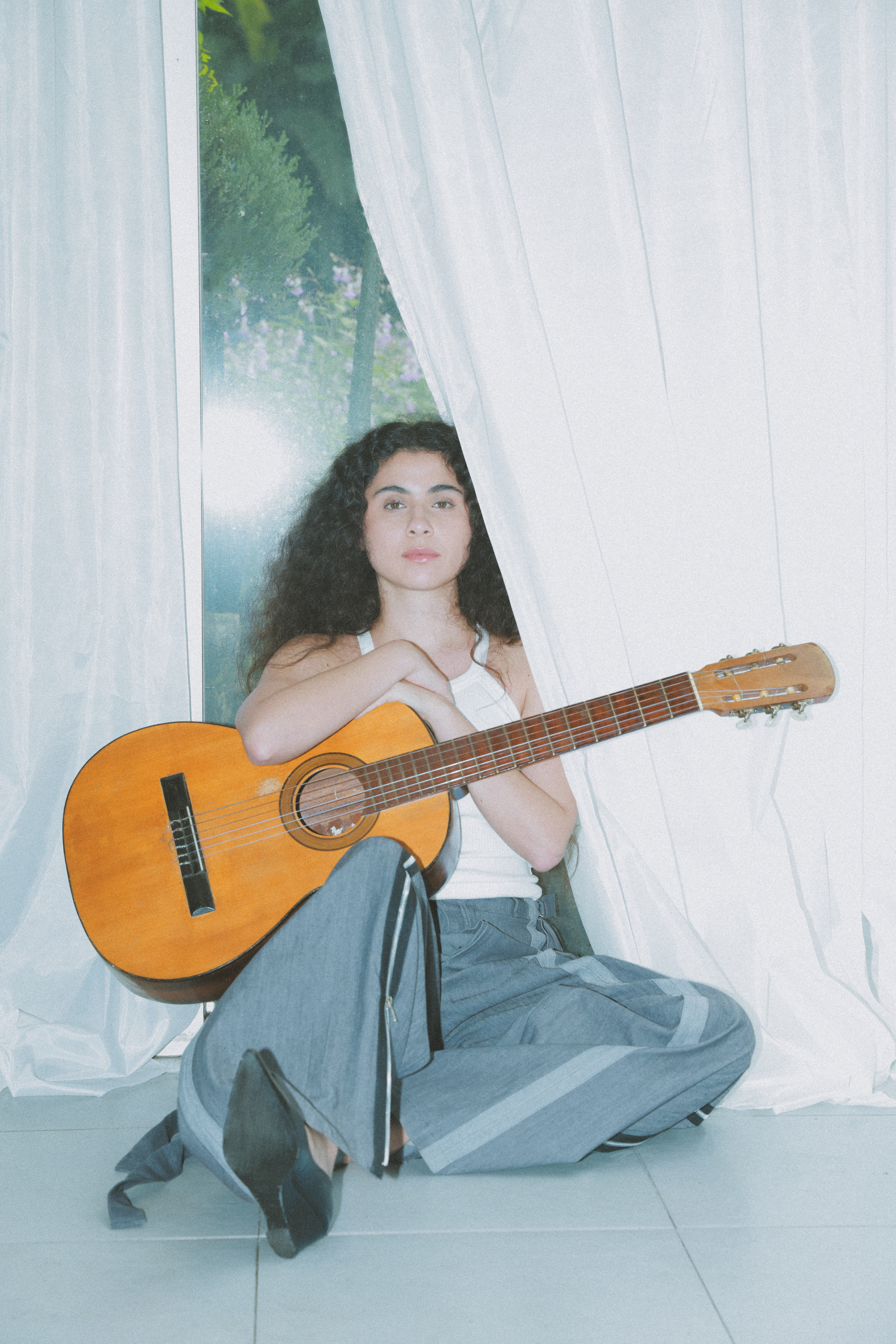Silvana Estrada sitting on the floor against a window holder her guitar.