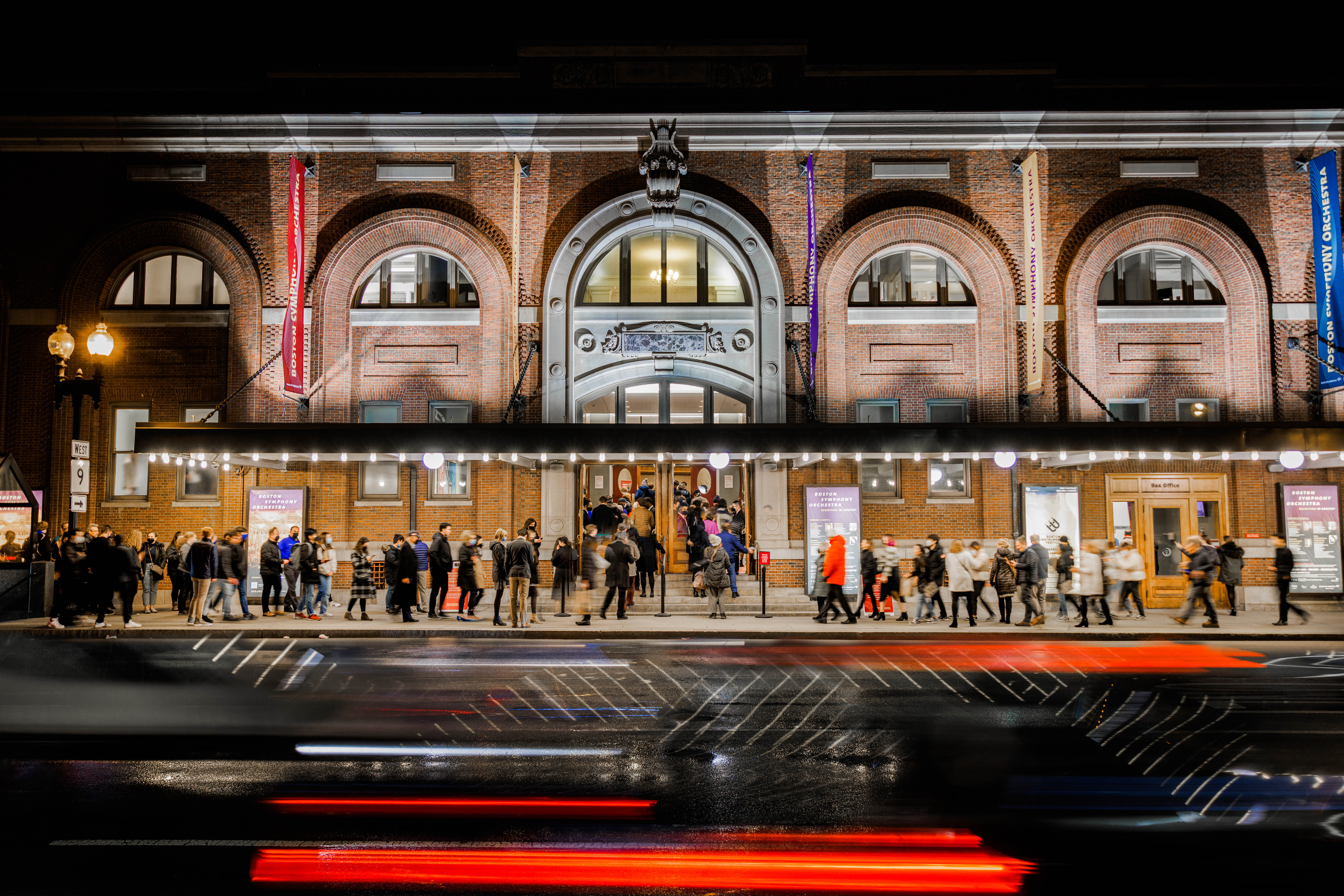 Audiences line up outside of Symphony Hall before a concert, with a blur of car headlights rushing past on the street