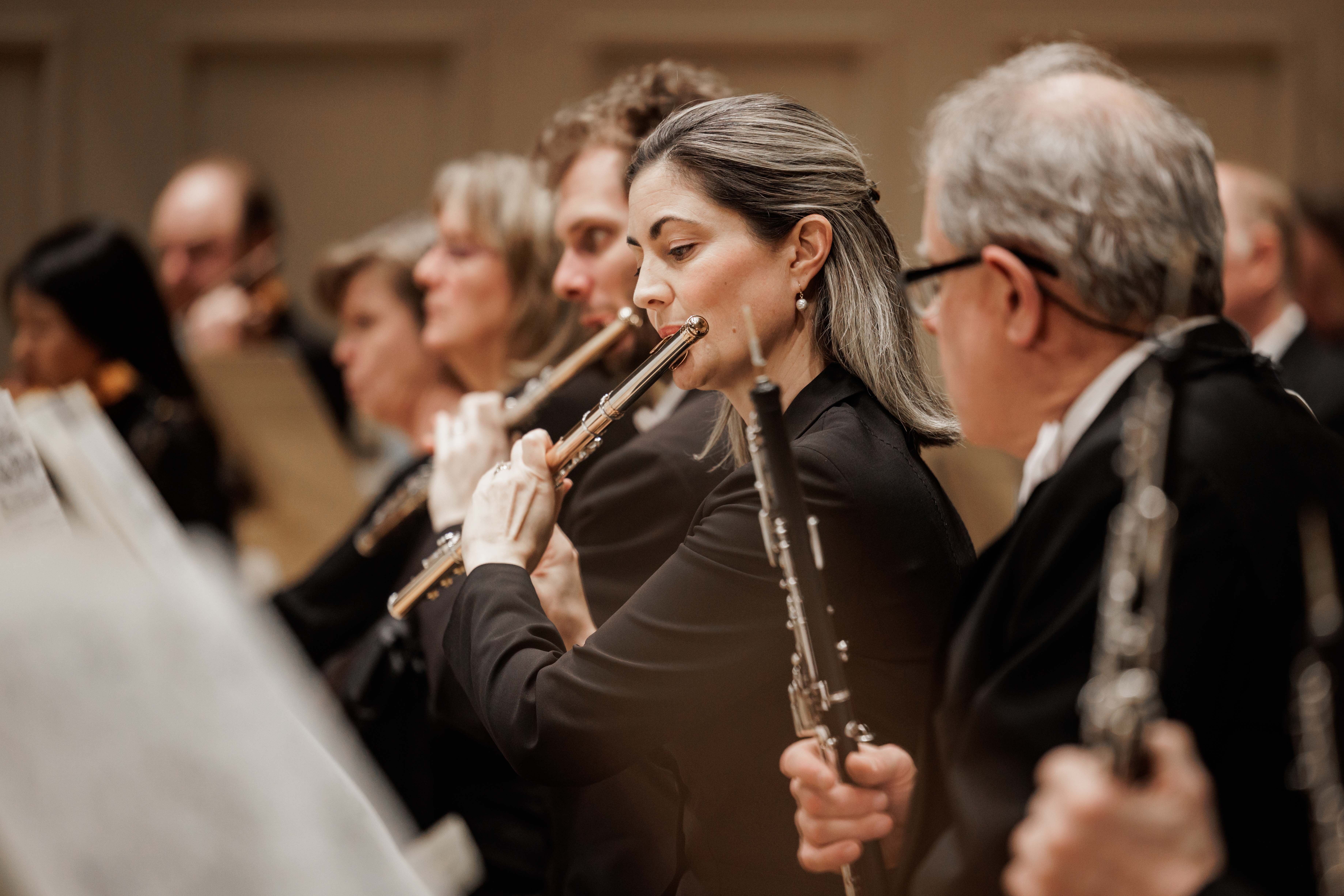 A woman with dirty-blonde hair plays the flute, with other flutists playing next to her