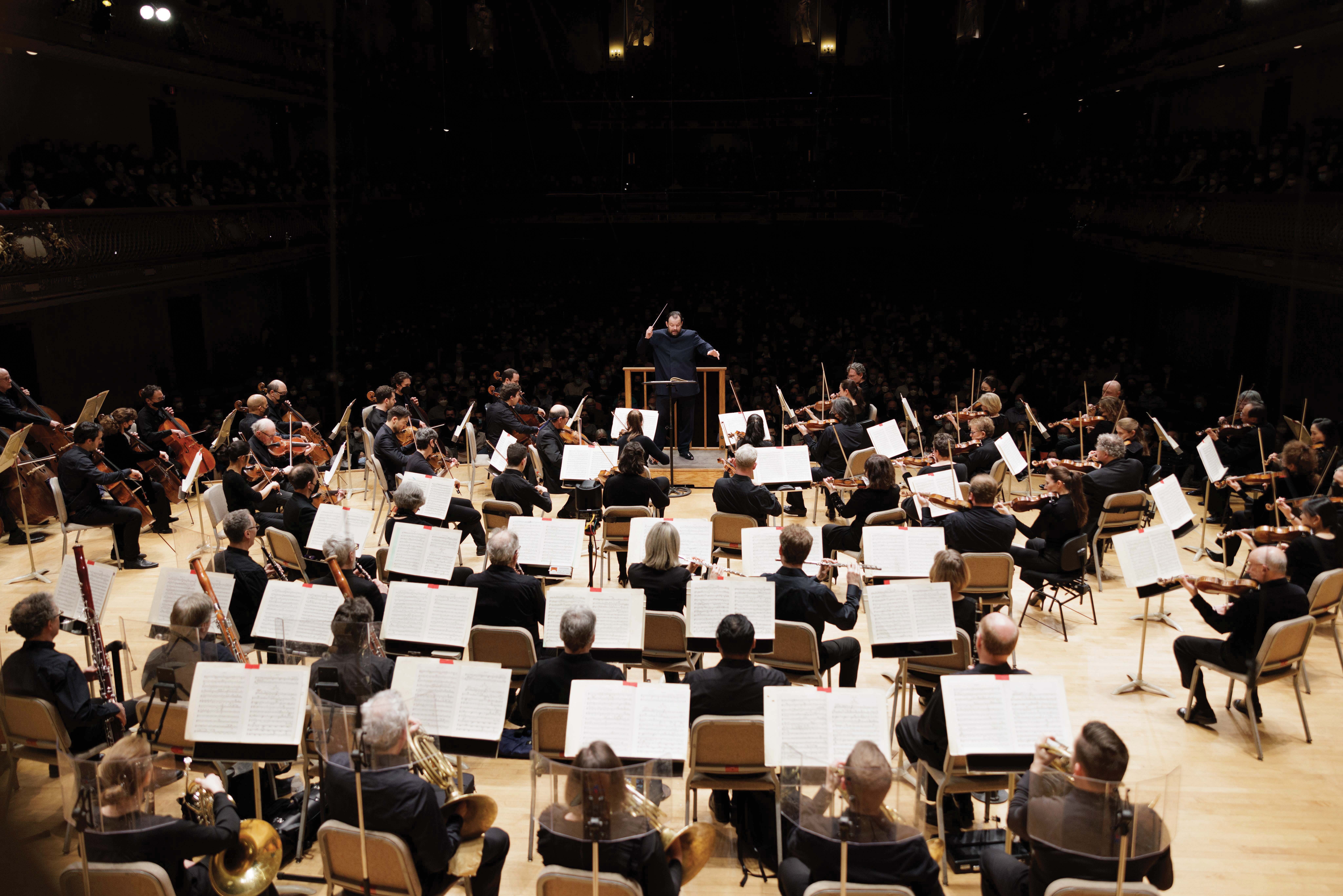 The BSO, all dressed in black, play on the Symphony Hall with Andris Nelsons conducting, the hall dark behind them