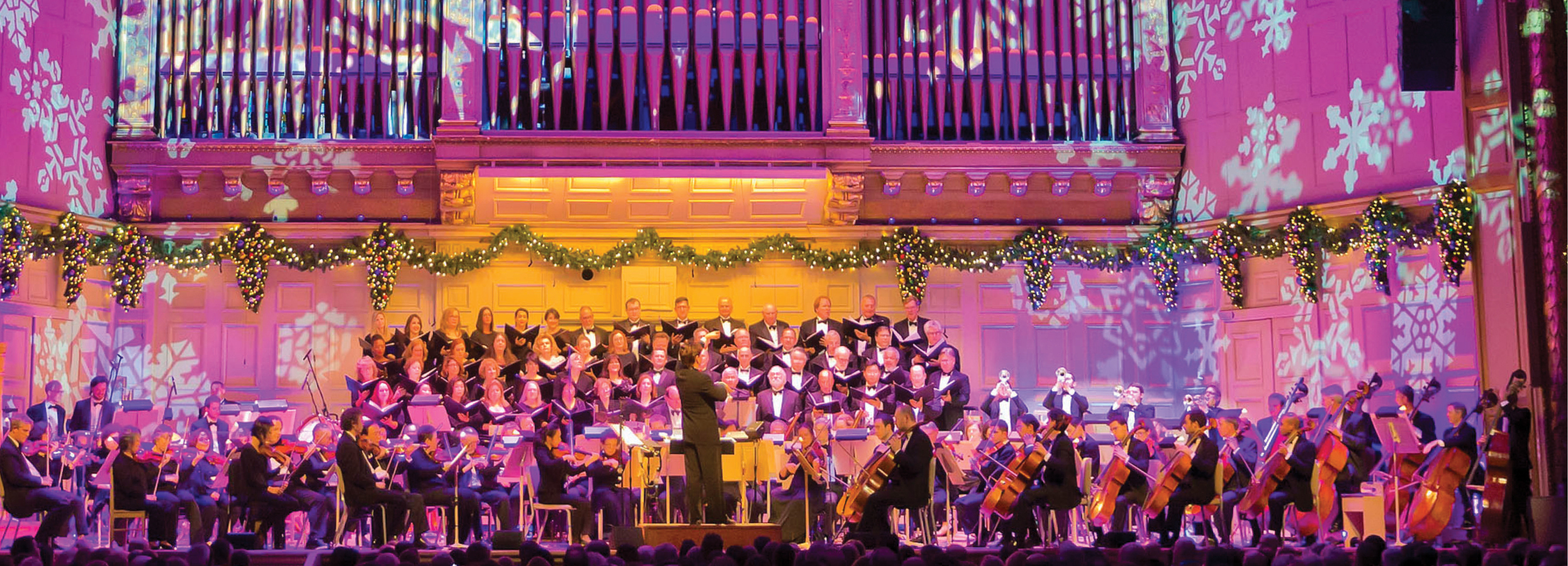 The Boston Pops and Tanglewood Festival Chorus perform on the Symphony Hall stage, which is lit with a bright purple light and projections of snowflakes