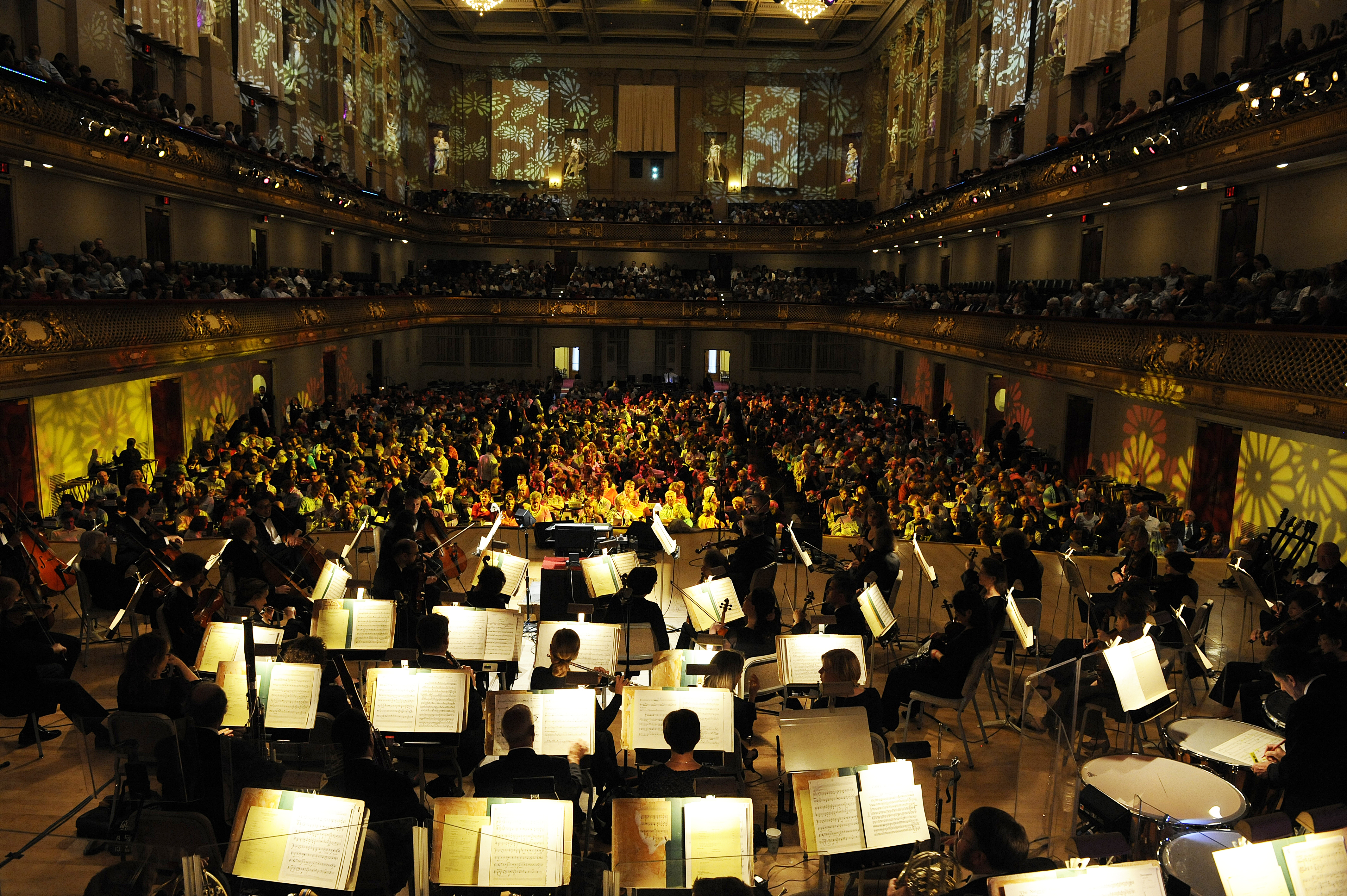 The Boston Pops tunes their instruments on a dark stage, their music stands lit up, as a large audience in Symphony Hall watches.