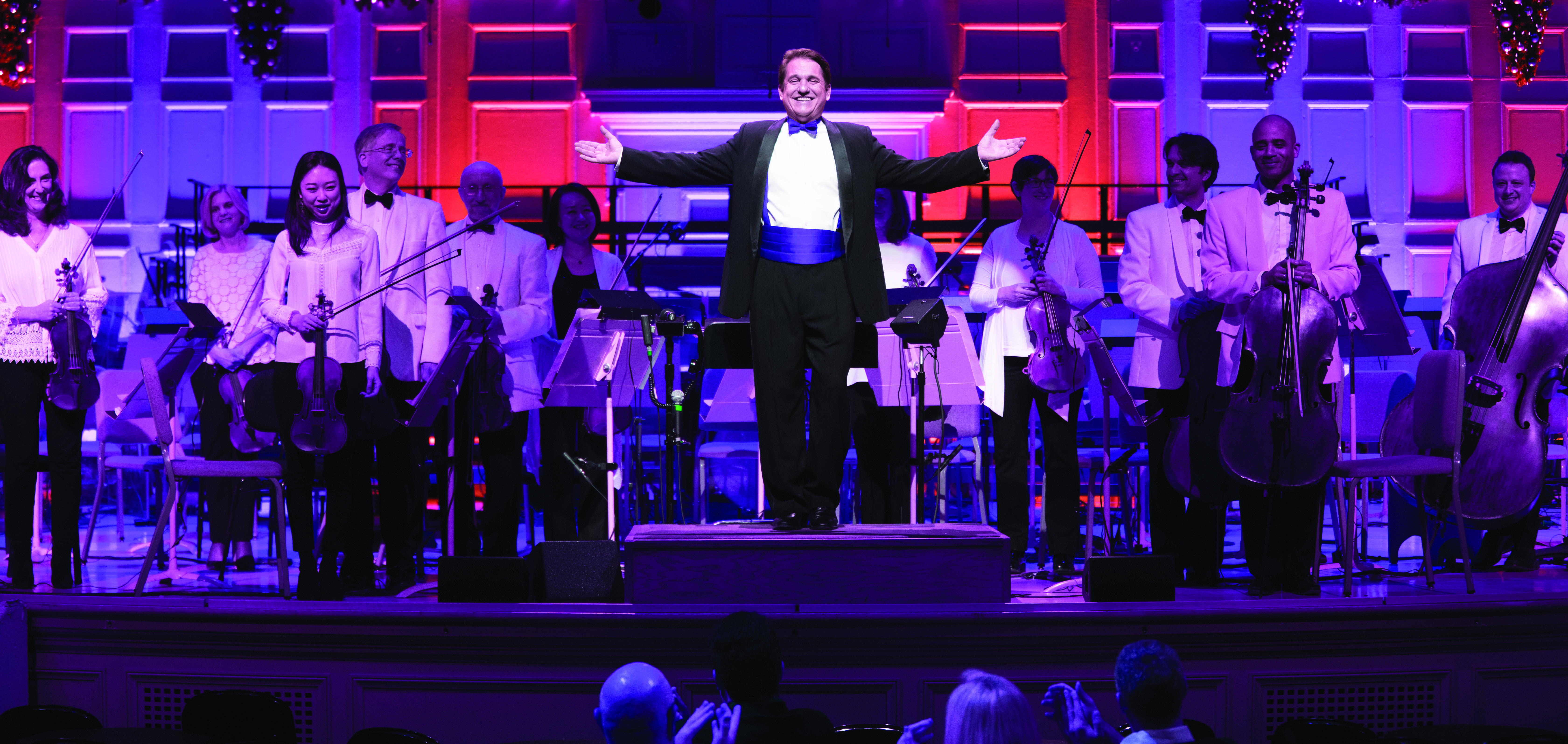 Keith Lockhart stands on the podium at Symphony Hall, smiling at the audience and opening his arms wide as the Boston Pops players stand behind him and smile