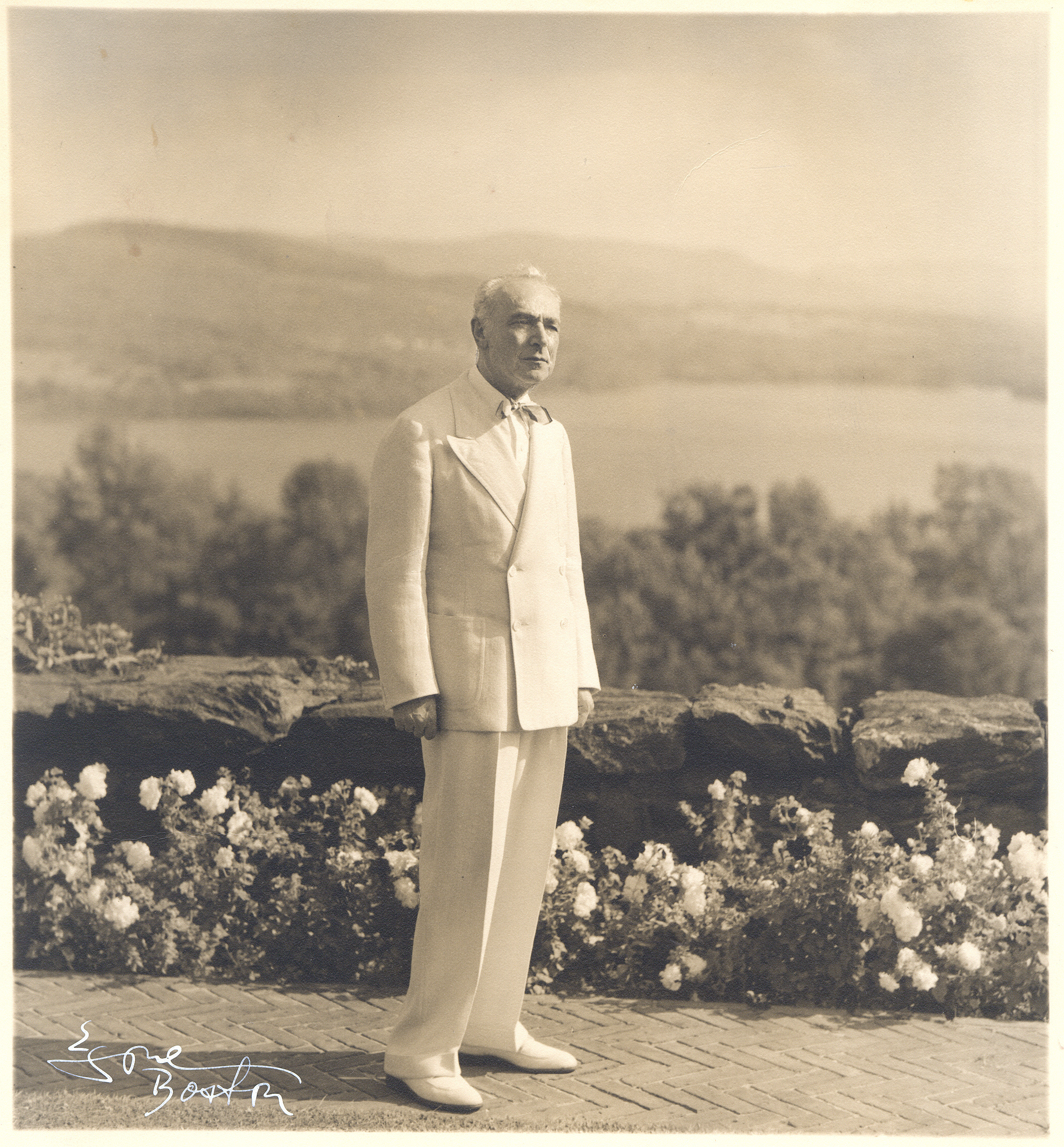 Serge Koussevitzky dressed in a white suit stands in front of a wall with a view of the Stockbridge Bowl lake behind him