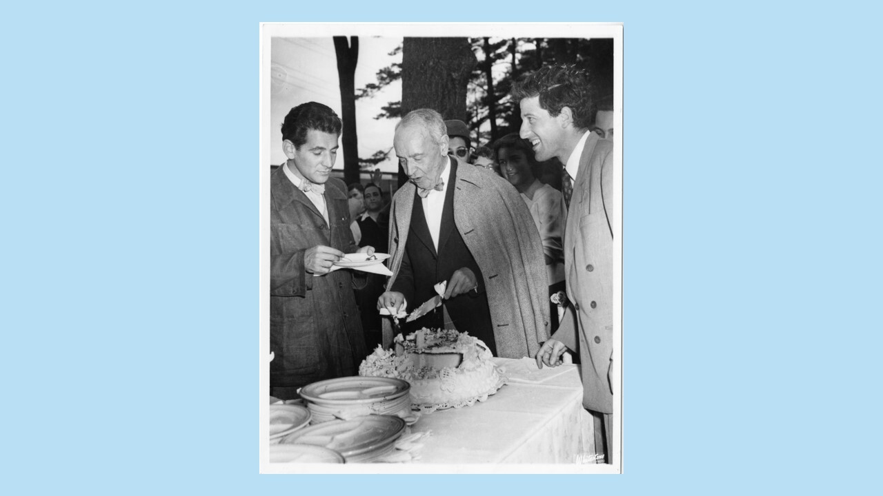 Leonard Bernstein and Lukas Foss stand on either side of Serge Koussevitzky as he cuts his birthday cake at Tanglewood ca 1946