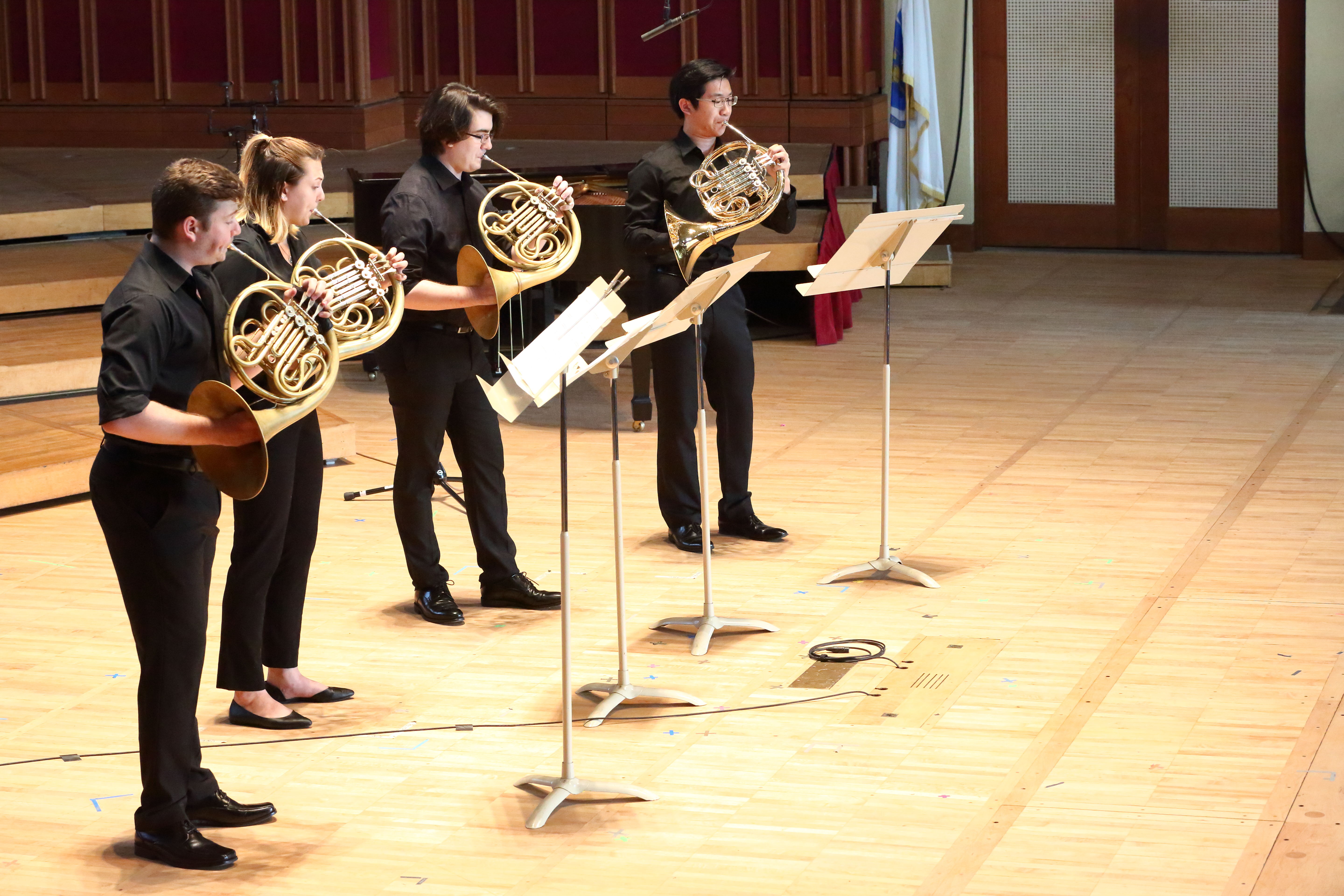 Four musicians dressed in black standing onstage playing French horns