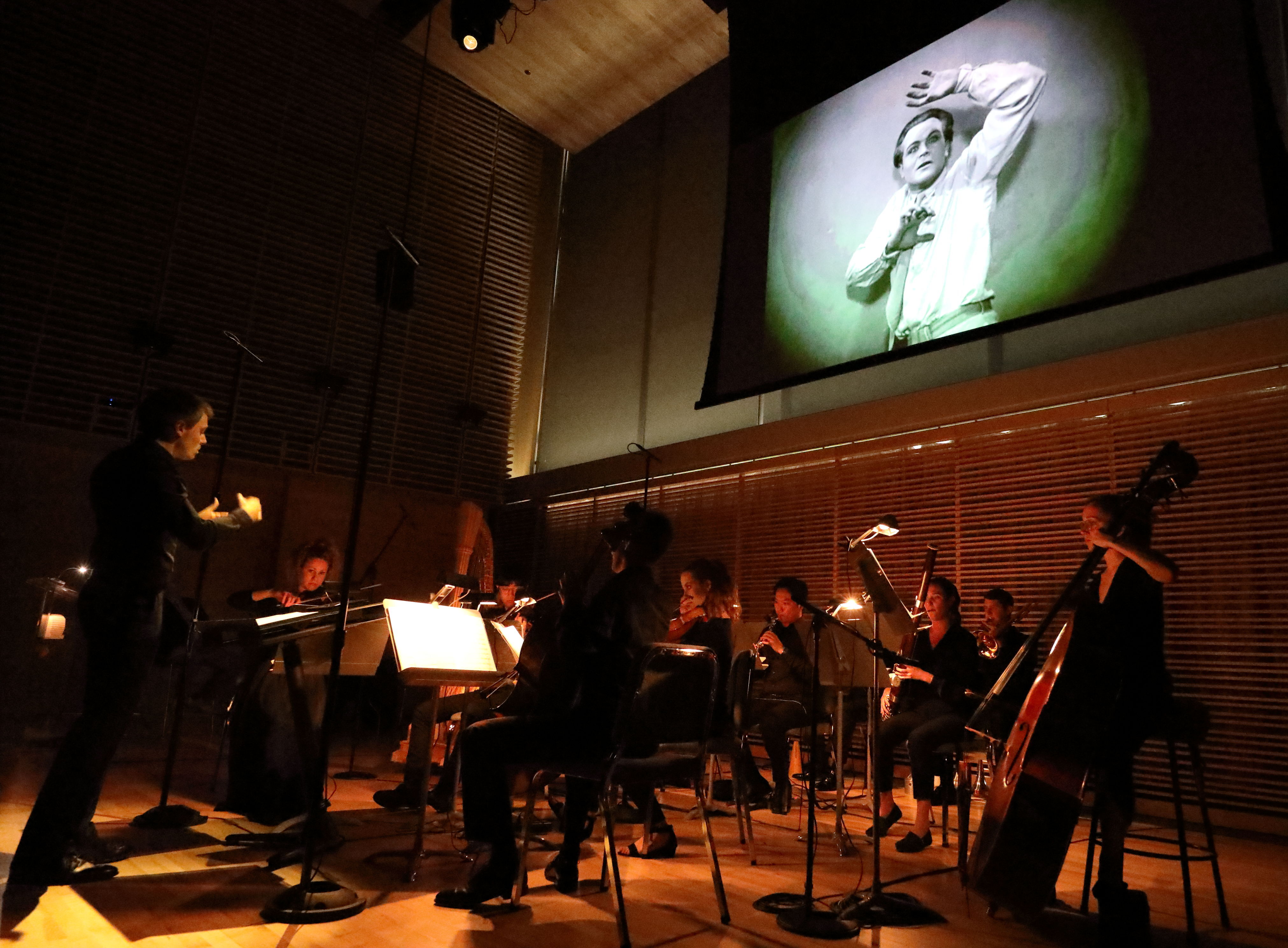A small orchestra playing beneath a screen with a black and white film projected on it.