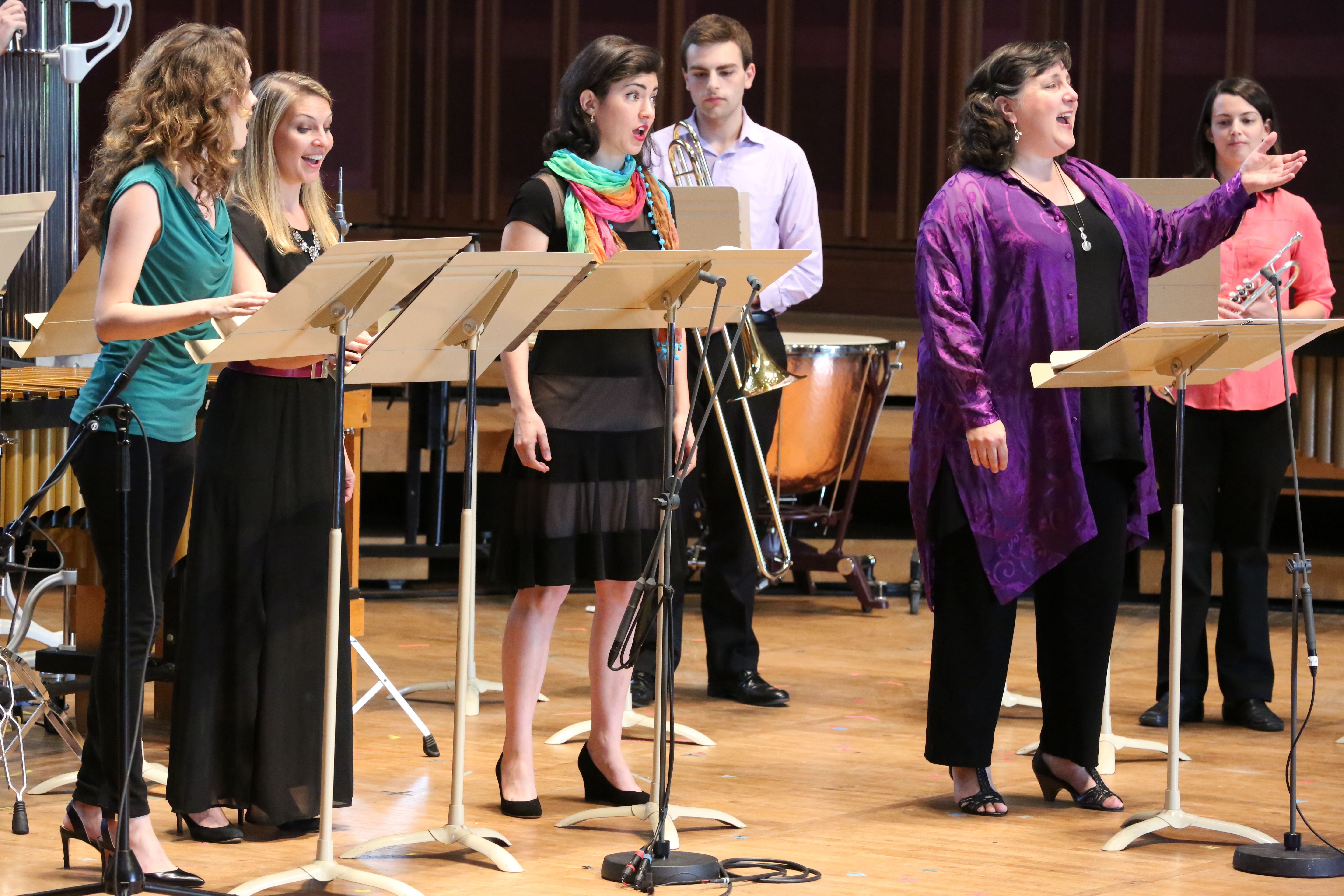 Four singers stand on stage behind music stands and sing, as two brass players stand behind them holding their instruments