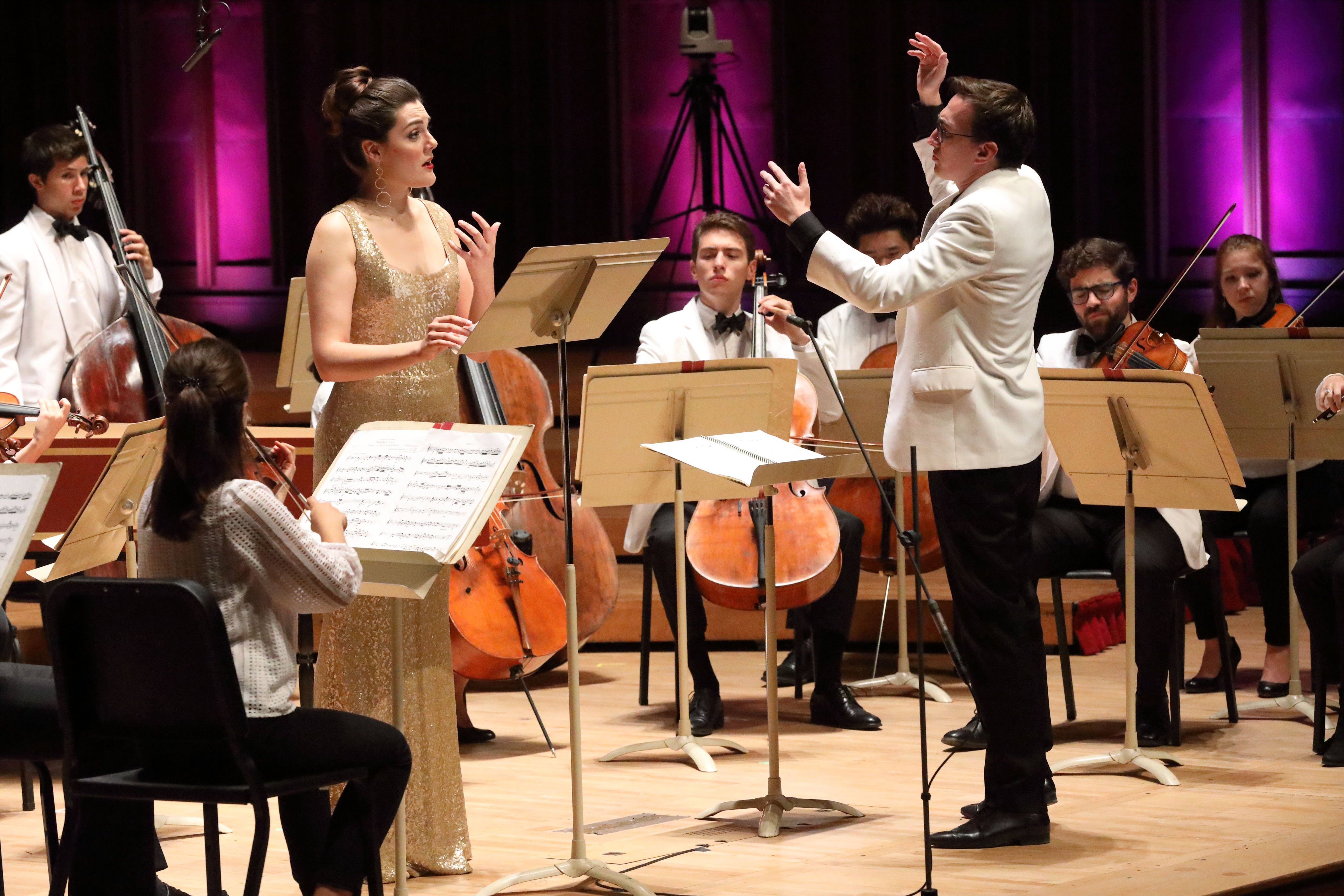 A vocalist performs onstage in a gold dress with string musicians and a conductor.