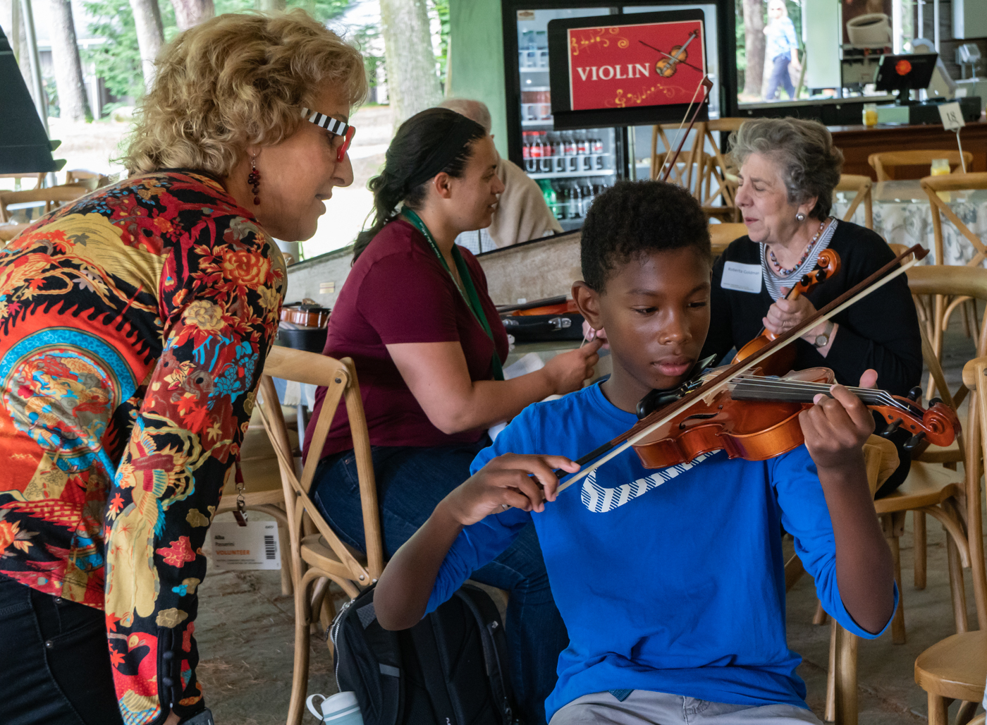 A woman in a colorful patterned shirt looks at a young boy's finger positions as he plays the violin