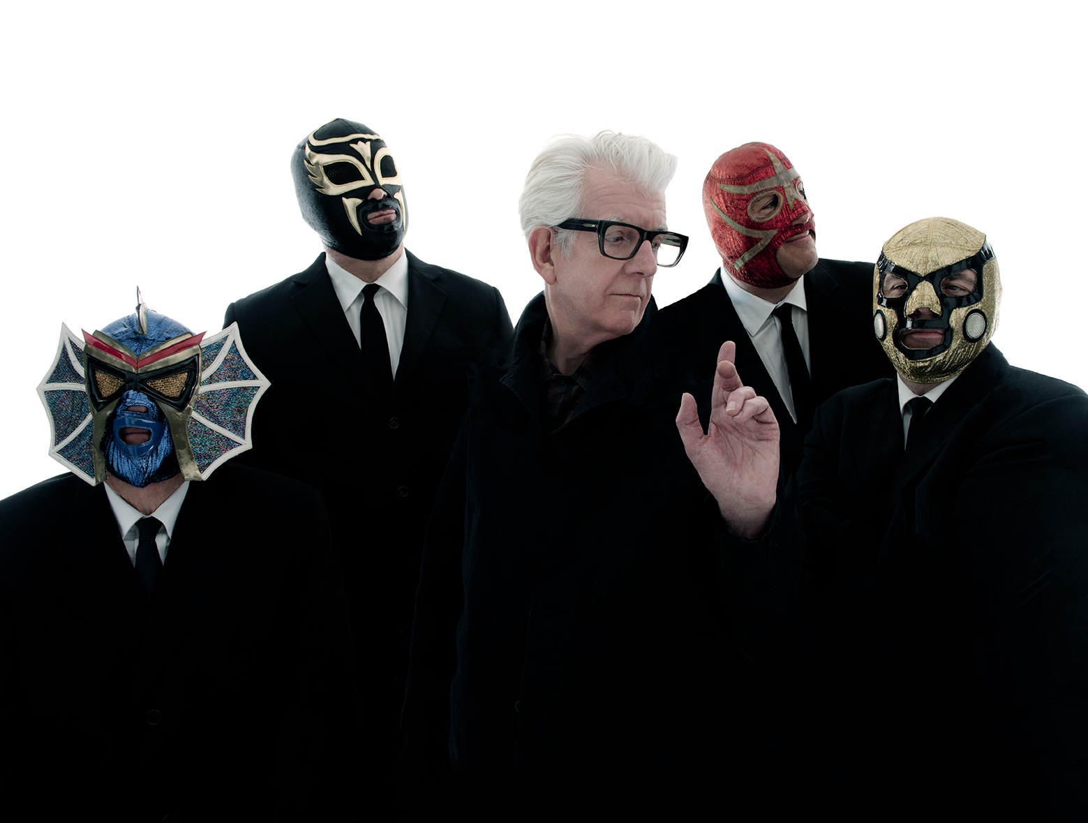 Nick Lowe wearing glasses and standing in front of four men dressed in suits and wearing face masks.