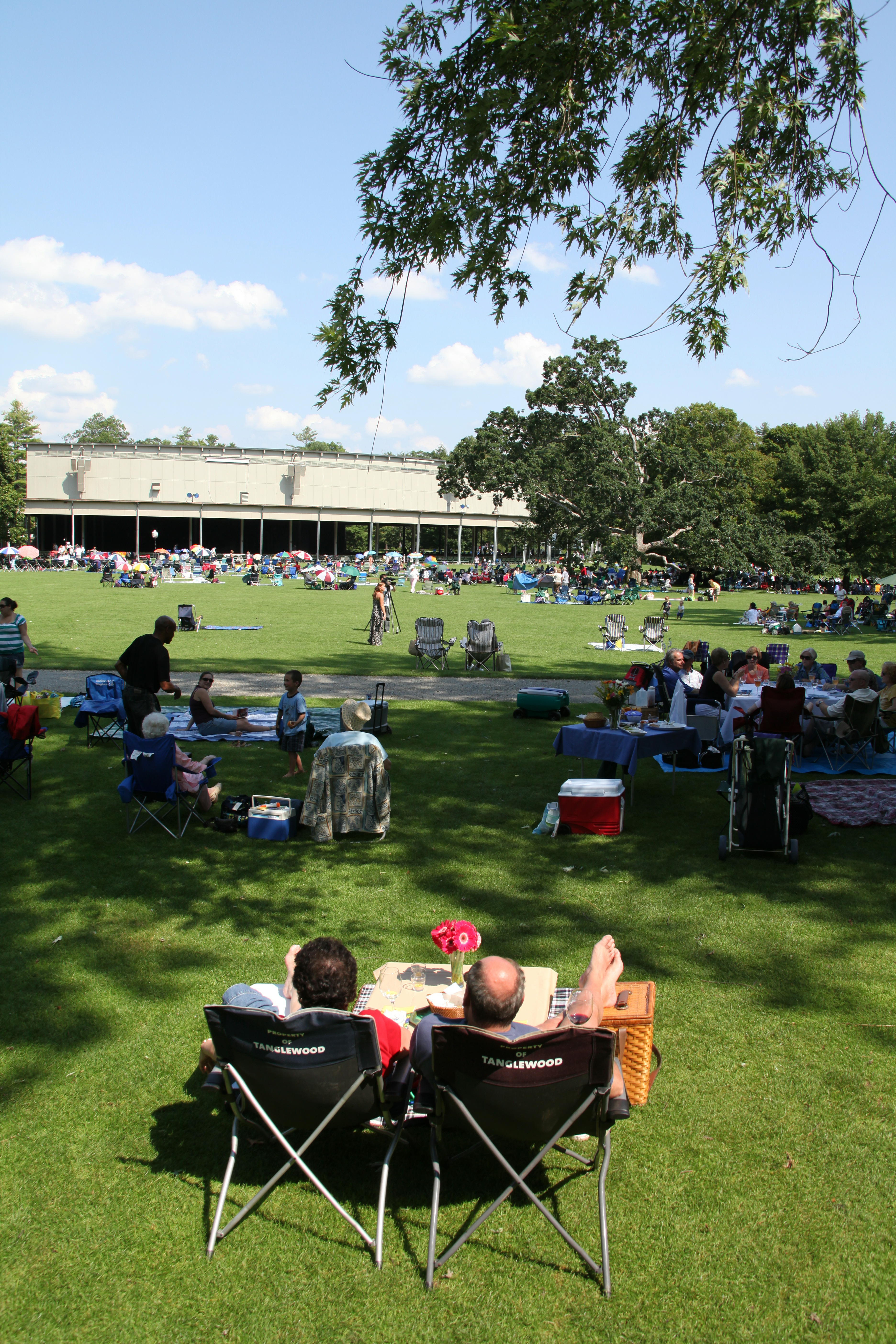 Two people sit in lawn chairs on the Tanglewood lawn behind a small table, facing the Koussevitzky Music Shed