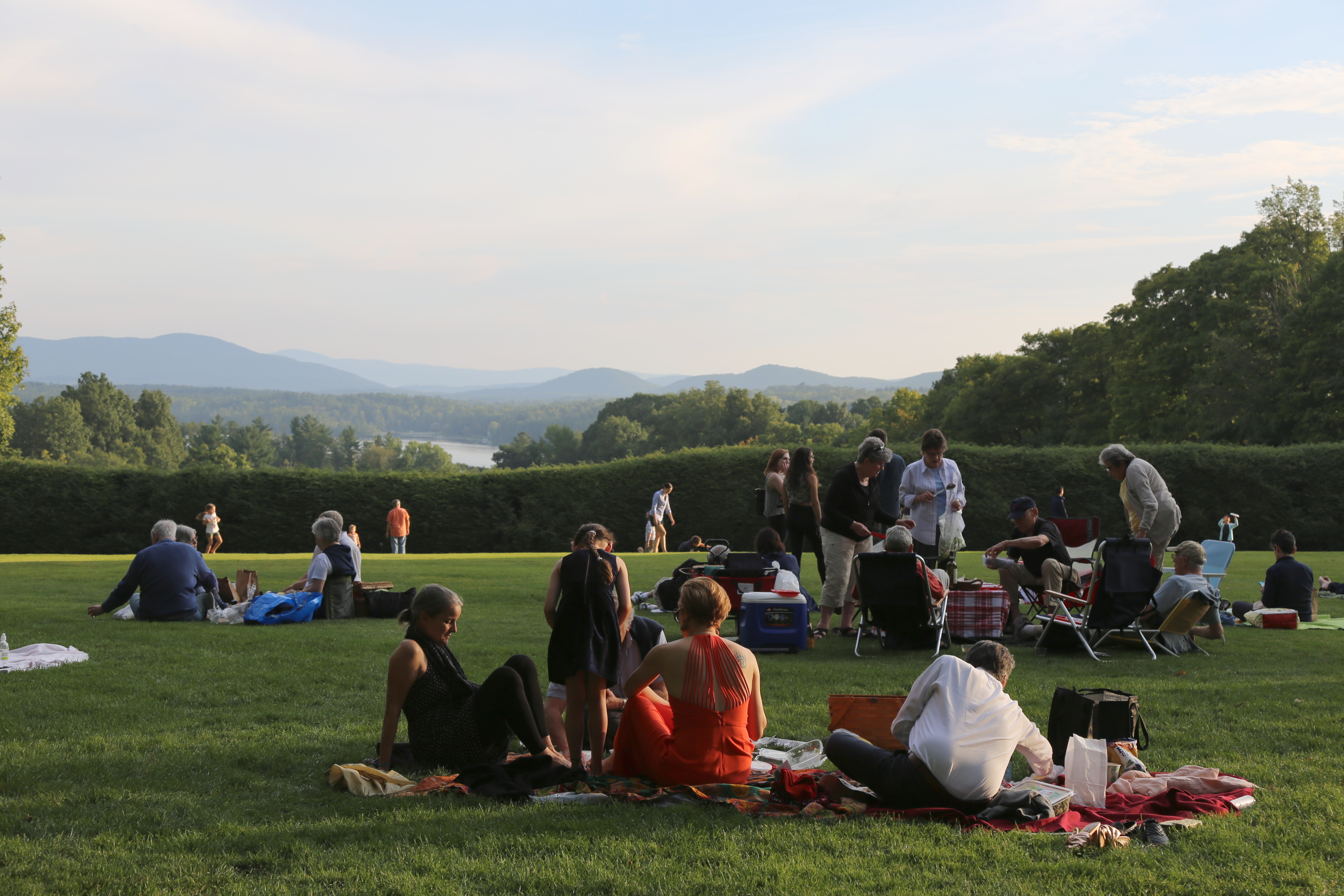 Three groups of people sit on green grass and picnic, with a view of the Stockbridge Bowl in the distance